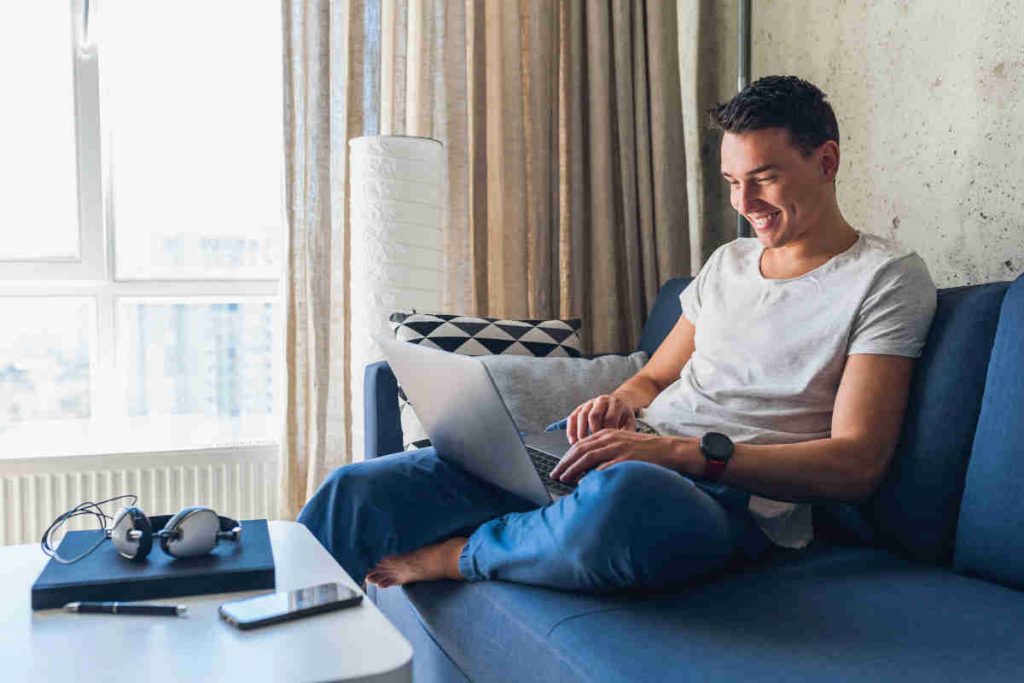 Giovane uomo seduto sul divano mentre naviga su internet con il suo laptop